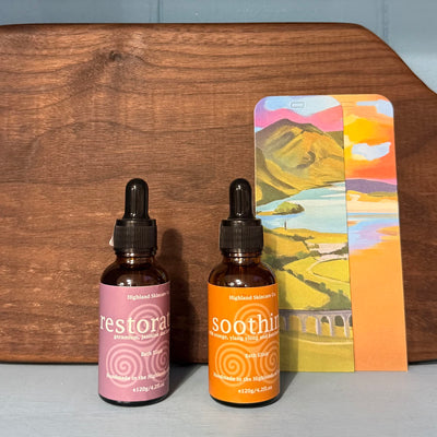 Two bottles of 'restore' and 'soothing' with a colorful landscape card on a wooden surface.