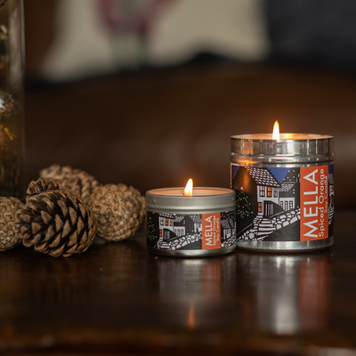 Two Mella candles with decorative labels on a wooden surface, surrounded by pine cones.
